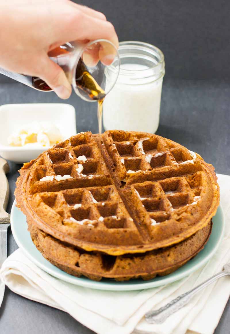 A human hand pours maple syrup over the top of two vegan sweet potato waffles.