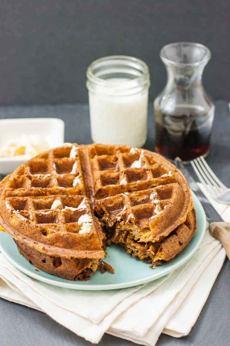 Oblique view of two vegan sweet potato waffles stacked on a teal-colored ceramic plate with a glass of dairy-free almond milk and tumbler of maple syrup in the background.