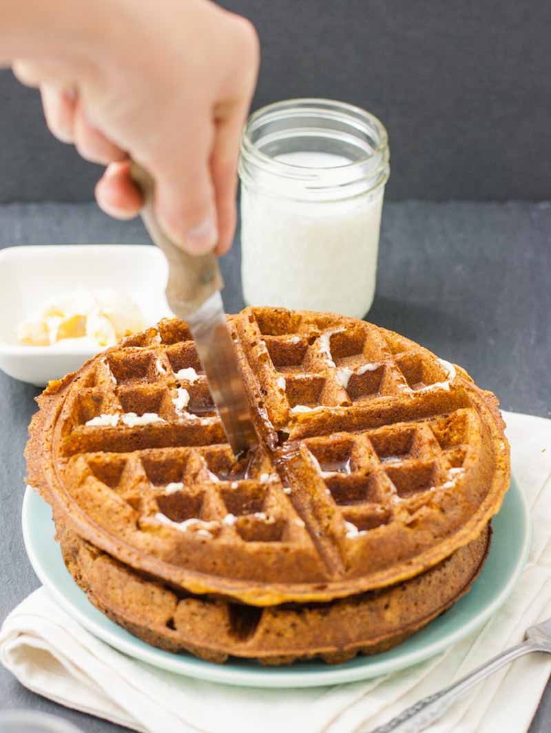 A hand cuts into a stack of two sweet potato waffles. A glass of almond milk is in the background.