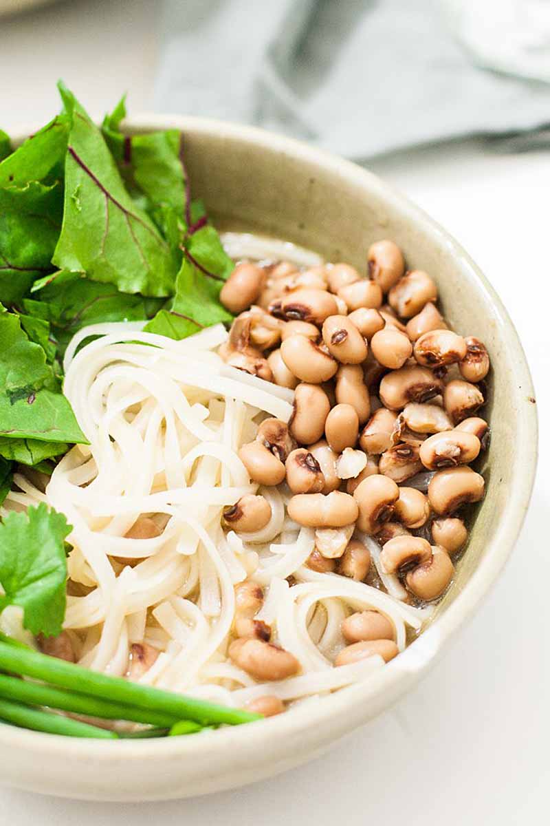 Close up of a bowl of black eyed pea noodle bowl that's gluten-free and vegan. Made for New Years with rice noodles that taste like ramen. Photo shows only right half the bowl on a white background.