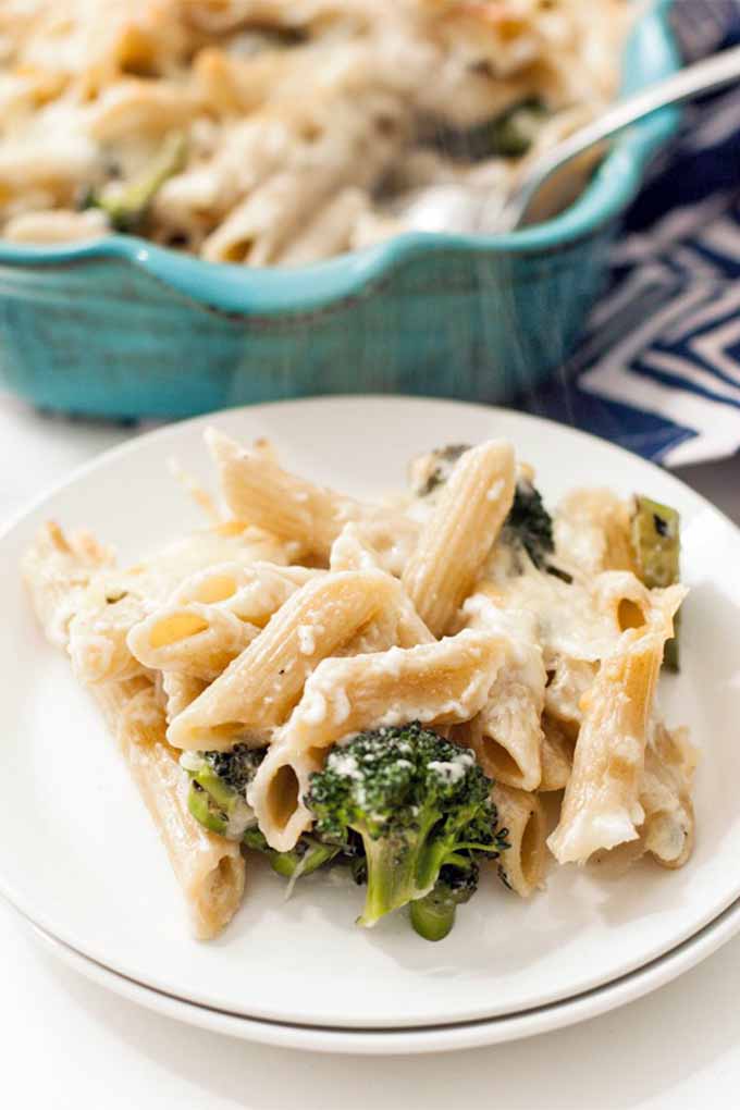 A plateful of penne pasta with melted cheese, bechamel, and broccoli, with a blue casserole dish in the background, on a white table with blue and white striped cloth.