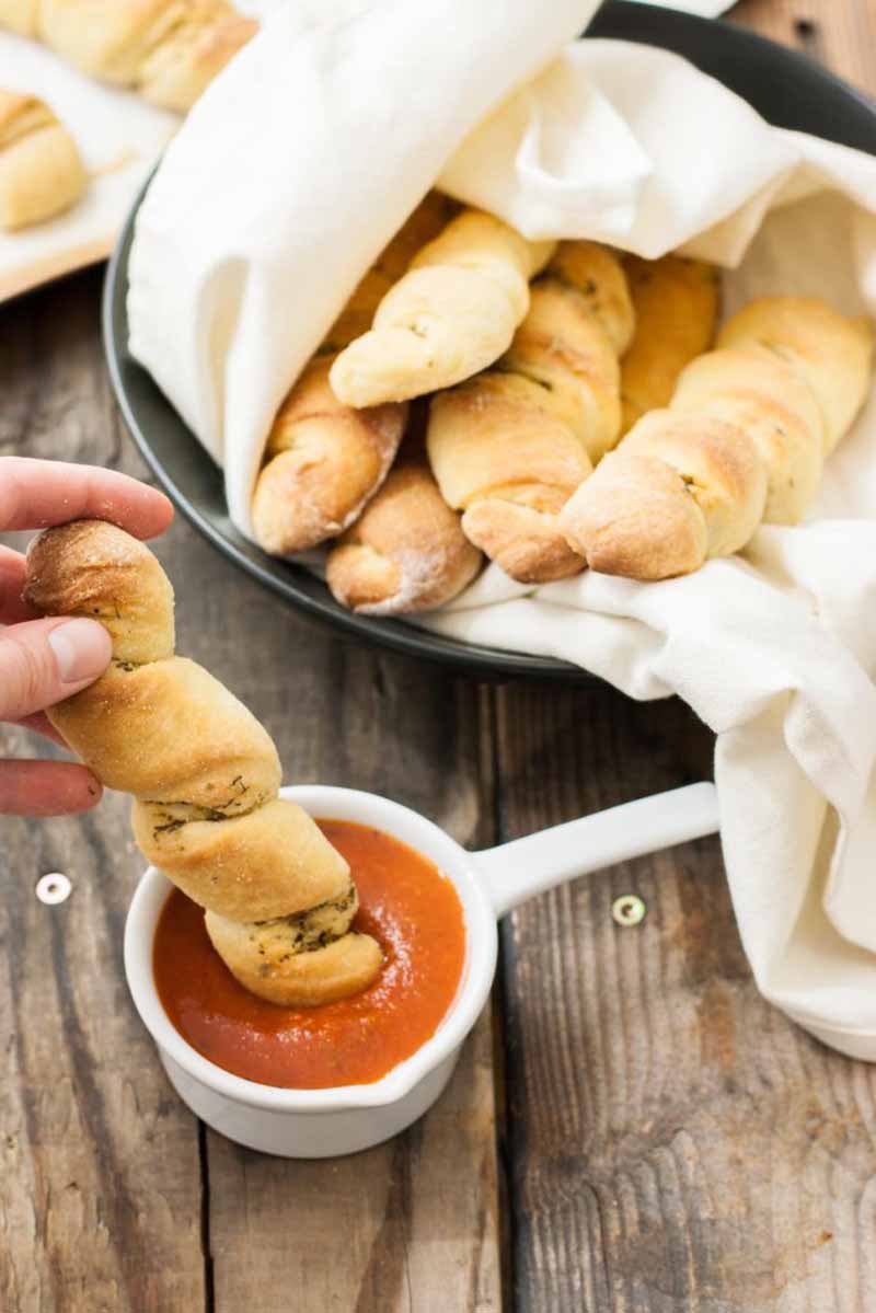 A human hand dips a no-knead Italian twisted breadstick into a white bowl containing red marina sauce. More breadsticks wrapped in a white towel are in the background.