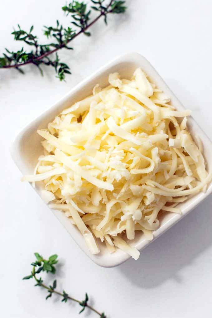 Top-down shot of a square dish of shredded Point Reyes Farmstead Toma cheese, on a white background with scattered sprigs of fresh thyme.