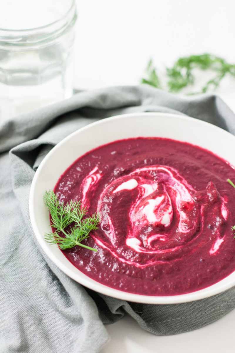 Oblique view of a white porcelain bowl full of purple roasted beet soup with swirls of a blonde cashew cream on top.