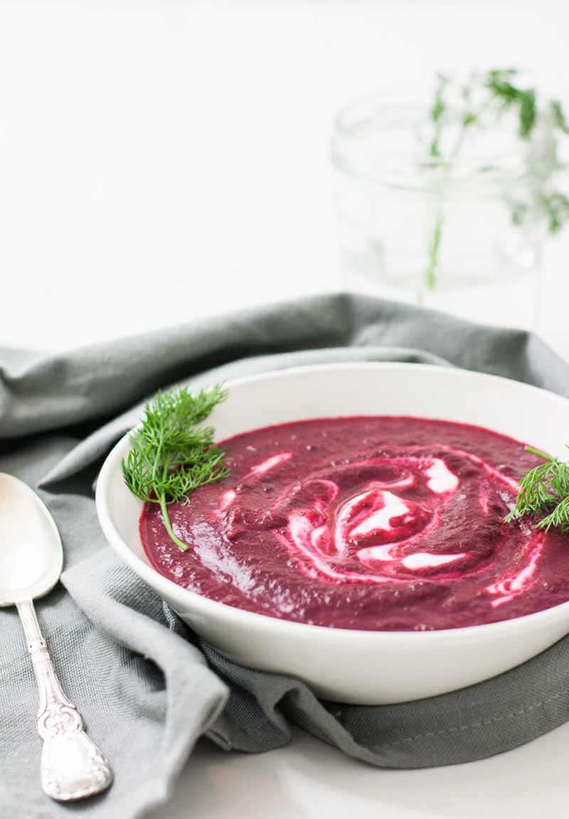 Oblique view of a purple roasted beet soup inside of white porcelain bowl with a gray cloth napkin and silver spoon to the left.