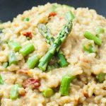 Closeup of a dark gray bowlful of beige-colored vegan asparagus risotto with oven-dried grape tomatoes.