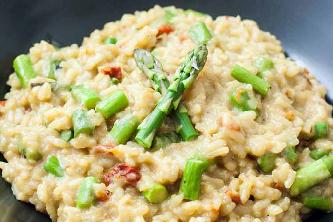 Closeup of a dark gray bowlful of beige-colored vegan asparagus risotto with oven-dried grape tomatoes.