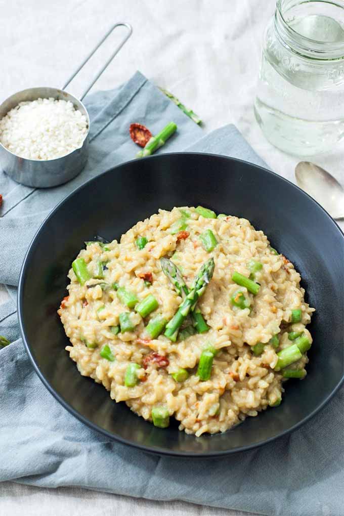 Vertical image of a black bowl of risotto with asparagus and oven-dried tomato, on a folded blue cloth with a metal measuring cup full of arborio rice, a piece of asparagus, an dried grape tomato, and a spoon.
