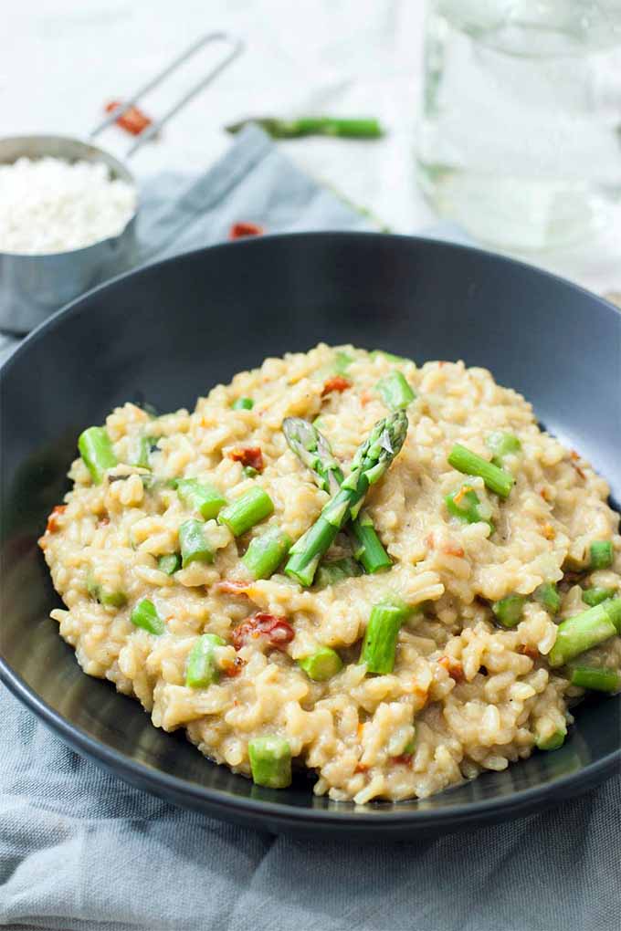 A large black ceramic bowlful of asparagus risotto with two spears arranged decoratively on top, flecks of dried grape tomato, and more vegetables and a measuring cup of arborio rice in the background, on a folded pale blue cloth.