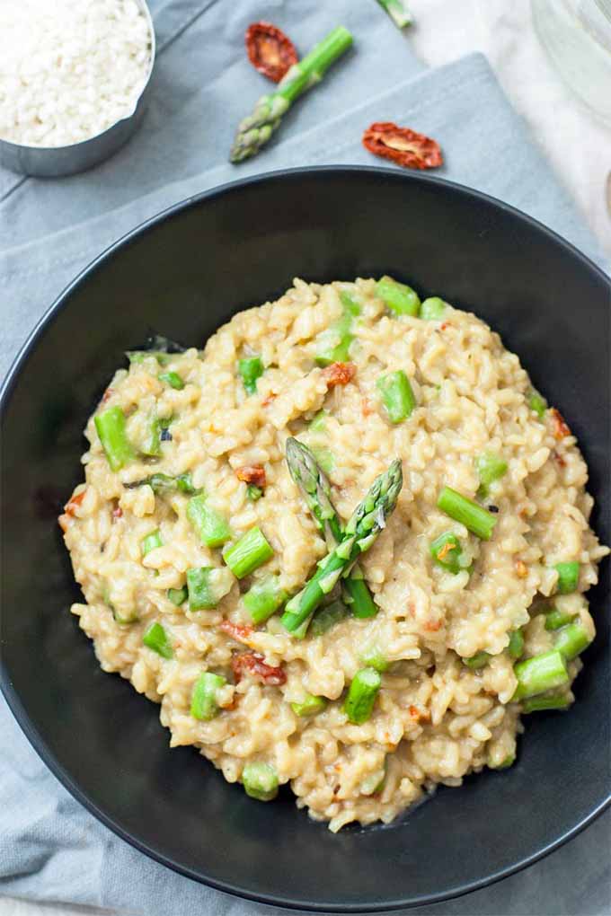 Top-down vertical image of a black bowl full of asparagus risotto, with pieces of dried tomato, on a folded light blue cloth with half of an asparagus spear, two dried tomatoes, and a silver metal measuring cup of white arborio rice.