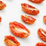 Closeup of three diagonal rows of dried grape tomatoes, with red crinkly edges and white centers, on white parchment paper.