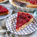 A slice of cranberry and brown butter custard tart is on a black and white patterned plate in the foreground, with another slice and the remainder of the dessert in a white ceramic pie dish in the background, with a metal and wood server, on a cloth surface with a fork.