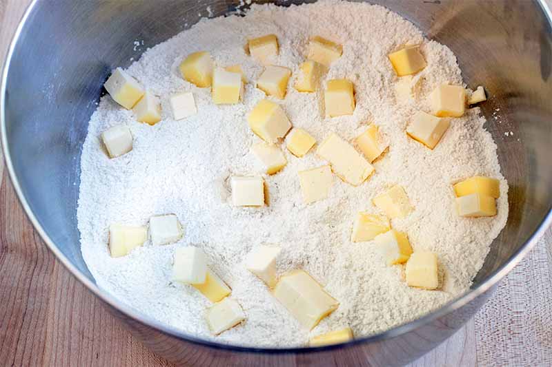 Cubes of chilled butter are scattered on top of a dry flour mixture in a stainless steel bowl.