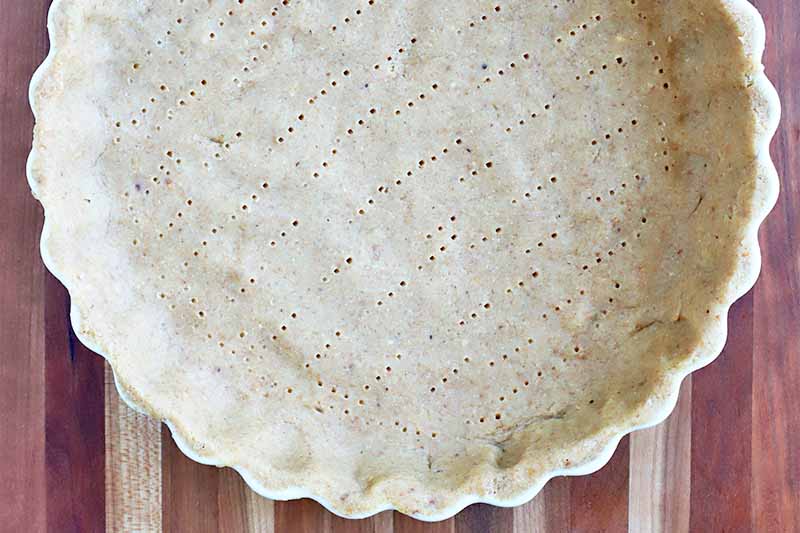 Top-down closely dropped shot of pie dough fitted into a ceramic baking dish, with an irregular pattern of holes all over the bottom, created with the tines of a fork.