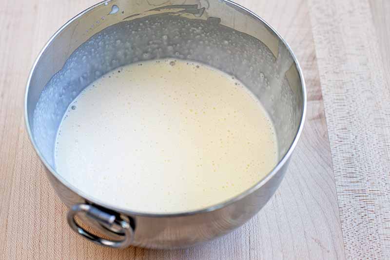 Cream and other liquid ingredients to make a batter fill a stainless steel mixing bowl with a ring handle, on a beige surface.