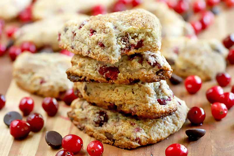 A stack of four scones is in the foreground, with scattered whole cranberries and chocolate chips, and more of the baked goods and fruit in shallow focus in the background, on a wood surface.