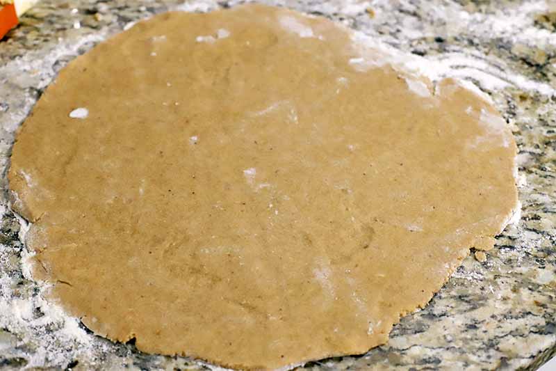 Homemade toasted hazelnut tart crust dough is rolled out into a shape that is roughly circular, on a floured granite countertop.