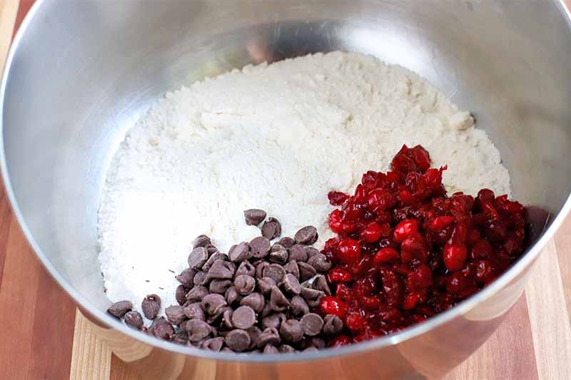 half of the area of the bottom of a stainless steel bowl is filled with flour, a quarter is filled with chocolate chips, and the remaining quarter is filled with chopped fresh cranberries, on a striped wood surface.