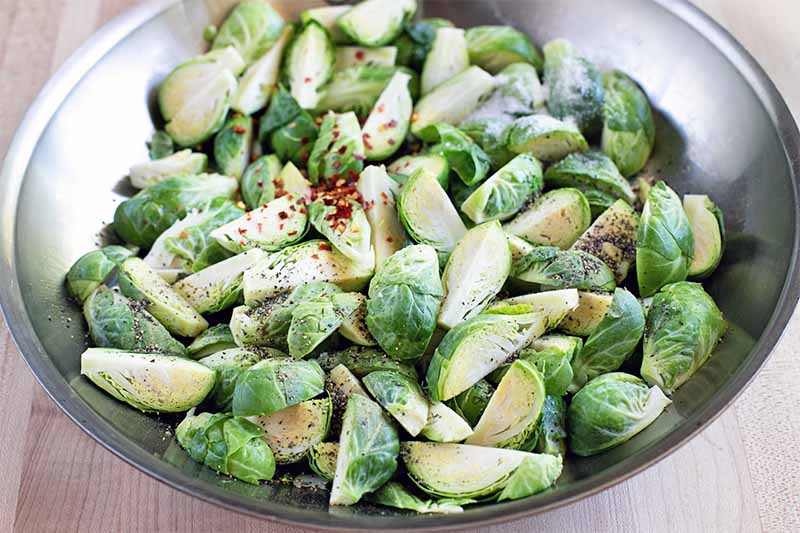 Cleaned and chopped Brussels sprouts with red chili flakes sprinkled on top, in a large stainless steel mixing bowl on a beige kitchen counter.