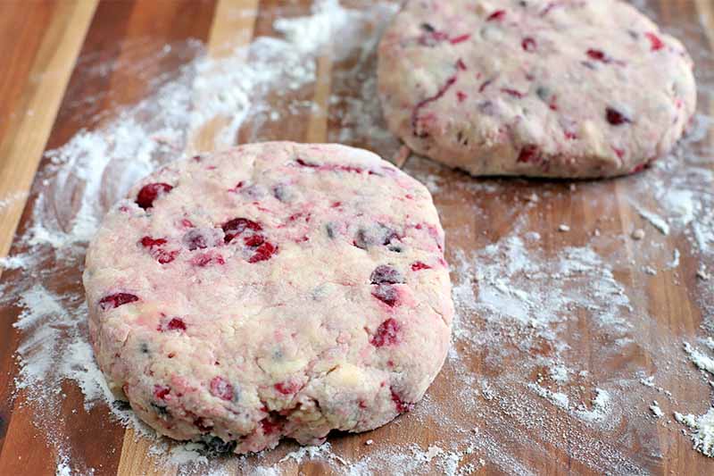 Two portions of cranberry and chocolate chip scone dough are portioned into round discs on a floured wood kitchen work surface.