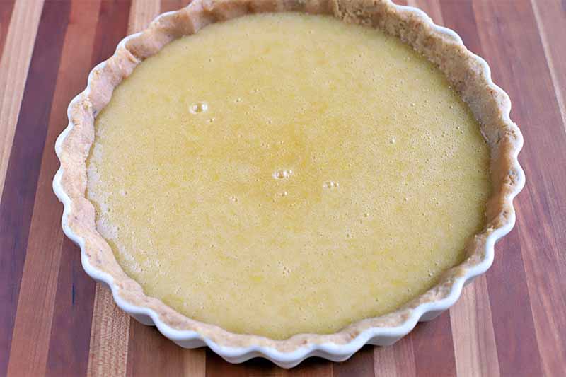 An uncooked brown butter egg custard mixture fills a par-baked crust in a white fluted ceramic tart pan, on a striped beige and brown surface.