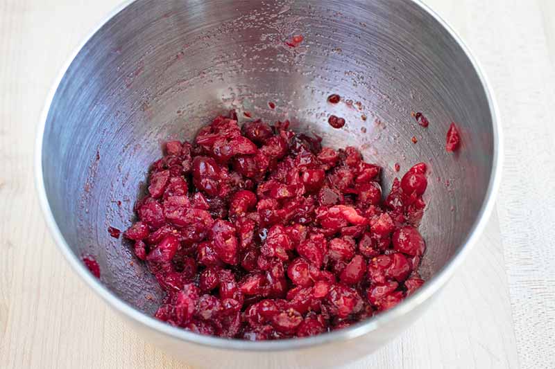 Chopped fresh cranberries in the bottom of a large stainless steel mixing bowl, on a beige surface.