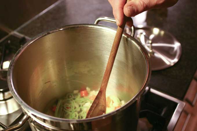 A hand stirs vegetables at the bottom of a tall metal stockpot with a wooden spoon.