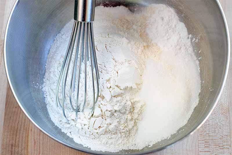 A metal whisk stirs a flour mixture for baking in a large stainless steel bowl, on a beige surface.