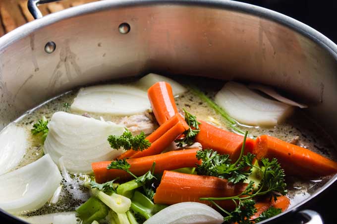 Closely cropped image of a stockpot filled with chopped carrots, onions, celery, herbs, and water.