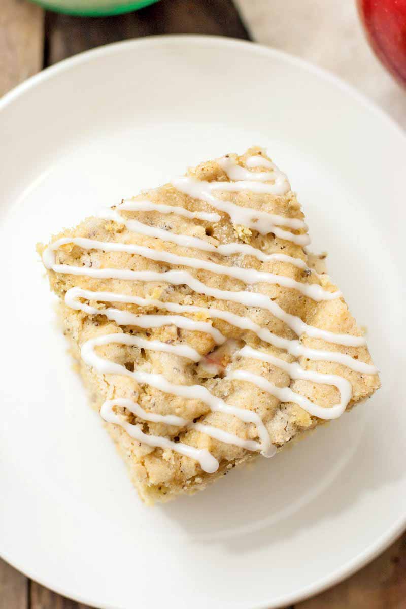 Overhead shot of a square piece of homemade vegan apple cake with a zig-zag drizzle of white icing on top, on a white plate, on a wood background.