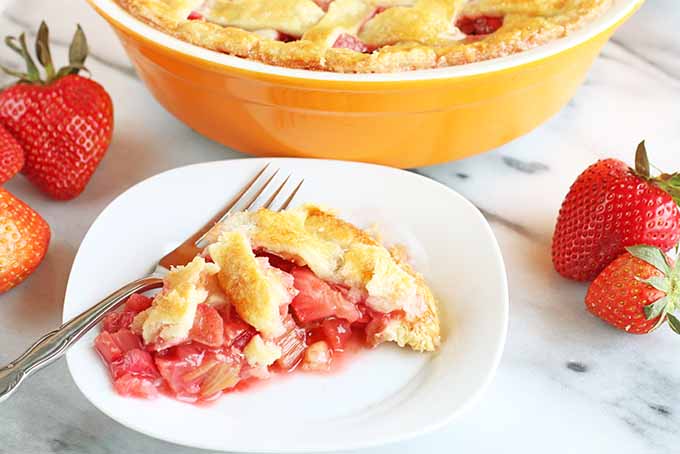 A piece of strawberry rhubarb pie is on a white square-shaped plate with rounded edges, alongside a fork, with the rest of the pie in a yellow-orange ceramic dish at the top of the frame, and several whole red strawberries with green tops scattered to the left and right on a white marble surface.