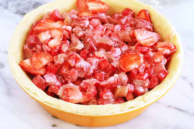 An orange ceramic pie dish is lined with dough rolled out thin to form a crust and filled with sliced strawberries and rhubarb, coated in cornstarch and sugar, on a marble surface.