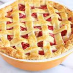 A just-baked strawberry rhubarb pie with a golden brown buttery lattice crust top, on a marble background.