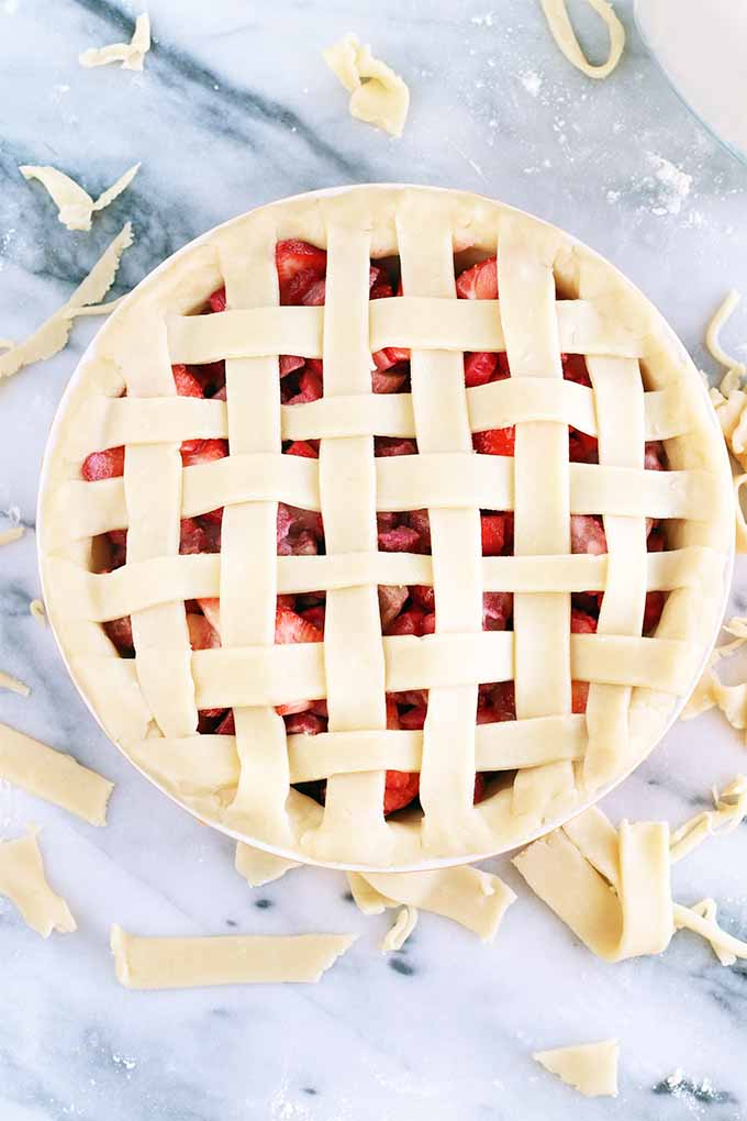 Top-down vertical shot of a homemade strawberry rhubarb pie with woven lattice crust that's ready to be baked in the oven, on a piece of gray and white marble with scattered strips and small pieces of rolled out dough.