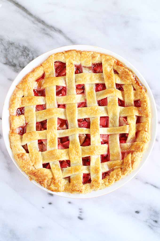 Top-down shot of a lattice-topped strawberry rhubarb pie, in a white ceramic pie dish on a gray and white marble background.