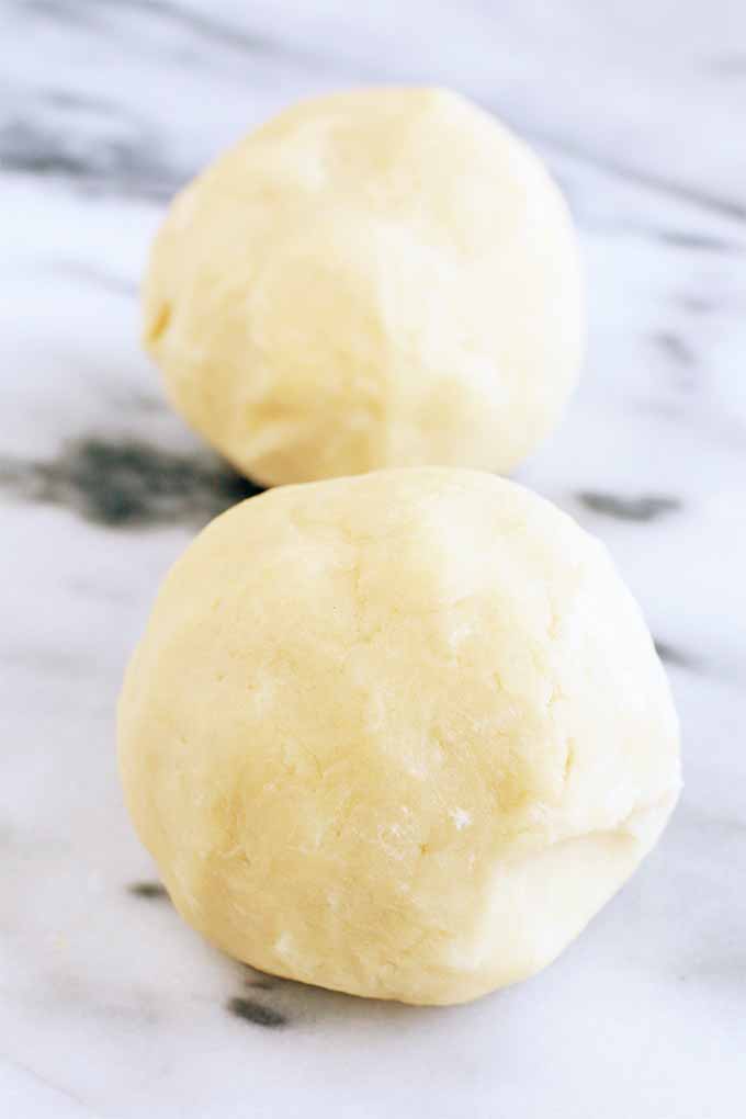 Vertical image of two equally-sized balls of pie dough, on a white marble surface with gray veins.