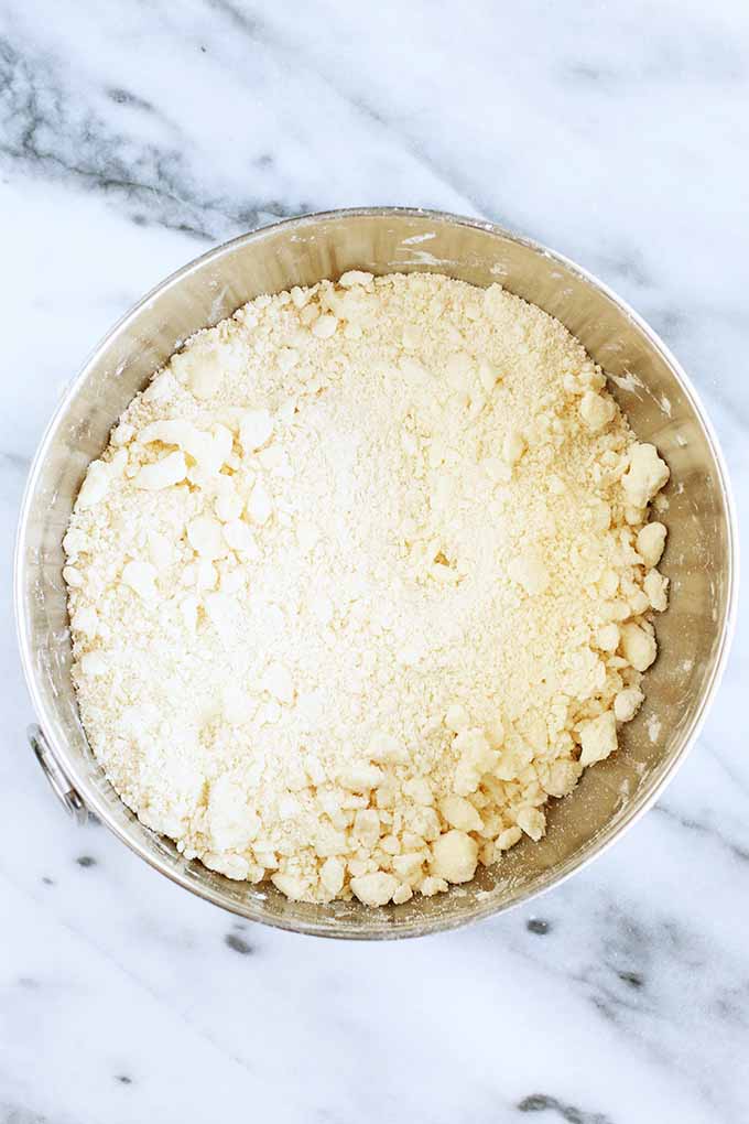 Top-down view of a stainless steel mixer bowl filled with a crumbly mixture of butter cut into flour to make a pie crust, on a gray and white marble background.