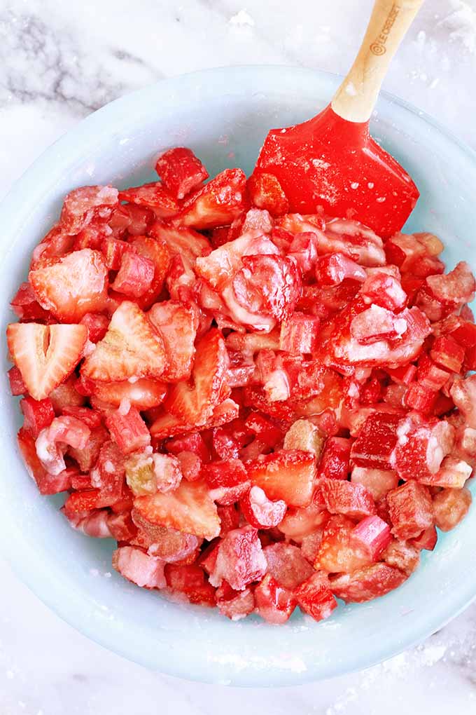 Top-down shot of a pastel blue mixing bowl filled with a mixture of sliced strawberries and rhubarb, sugar, and cornstarchm, being stirred with a red rubber Le Creuset spatula with a wooden handle, on a gray and white marble background.