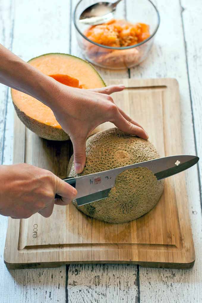 Closeup of a woman's hands, peeling a cantaloupe on a wooden cutting board with a chef's knife, with a glass bowl to hold the discarded rinds, on a white painted wood background.