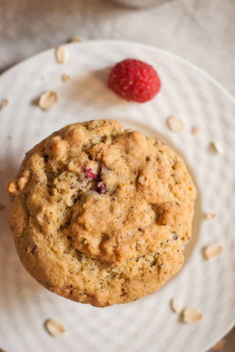 Top-down shot of a brown muffin on a plate, with a raspberry and scattered oats.