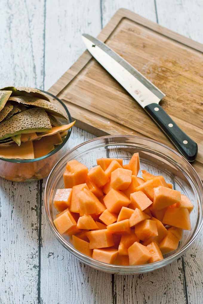 A large glass bowl of cantaloupe pieces sits in the foreground, with a smaller bowl of discarded rinds, a wood cutting board, and a chef's knife, on a white wooden table with boards oriented vertically in the background.