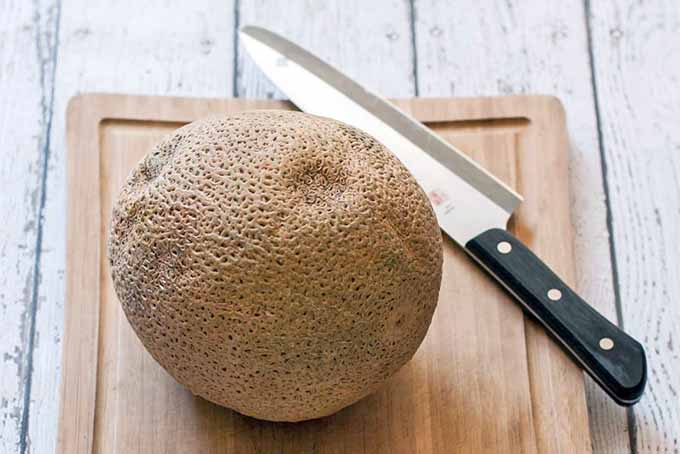 A whole cantaloupe rests on a wooden cutting board beside a chef's knife with a black handle, on a white wood table.