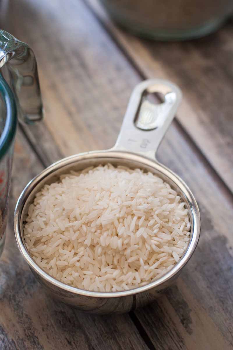 Oblique closeup view of uncooked white rice in a small metal measuring cup on a wood surface, with a glass pitcher-style liquid measure to the left of the frame