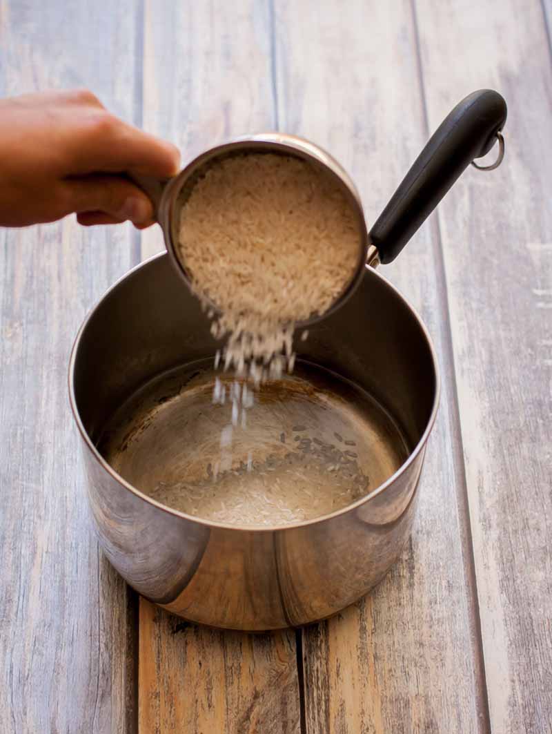 A hand pours uncooked white rice grains into an empty saucepan, on a wood surface.