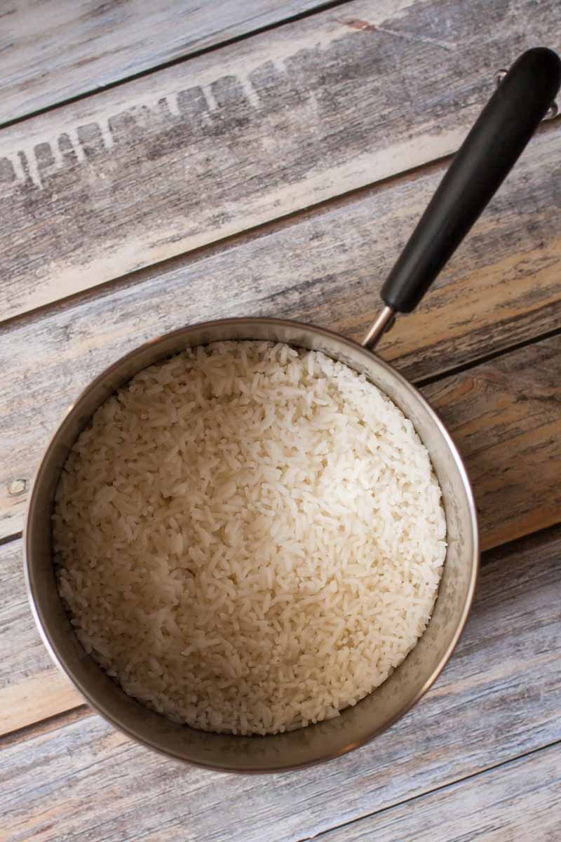 Top-down view of a metal saucepan of cooked, unfluffed rice sitting a rustic wooden tabletop. The pan has a black handle, and the image is in partial shadow.