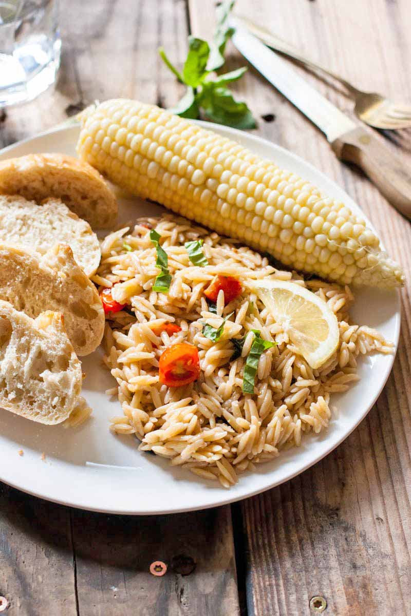 An ear of corn, pieces of bread, and a serving of orzo pasta salad with tomatoes, basil, and lemon slices, o a white plate, with a knife and a sprig of basil in the background, on a brown unfinished wood table.