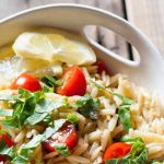 Closely cropped image of orzo pasta salad with halved cherry tomatoes, chopped basil, and slices of lemon, on a brown wood surface.