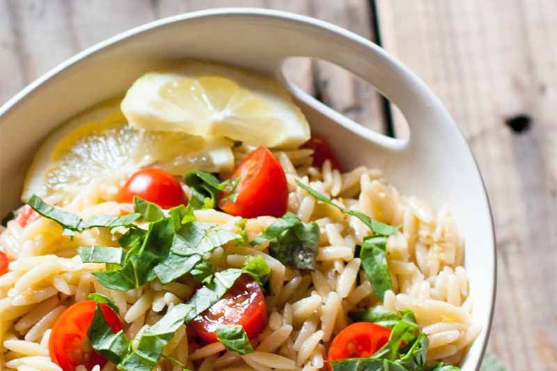 Closely cropped image of orzo pasta salad with halved cherry tomatoes, chopped basil, and slices of lemon, on a brown wood surface.