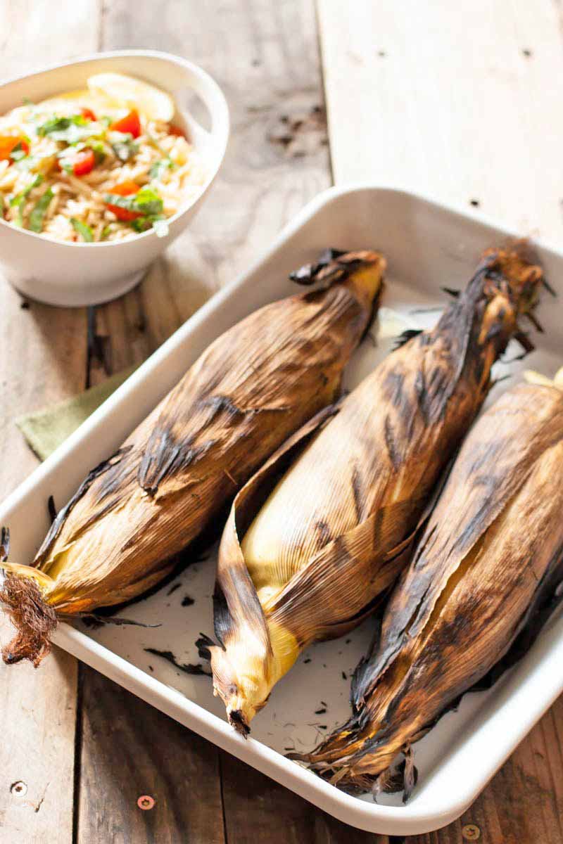 Three ears of grilled corn with the husks on in a rectangular ceramic baking dish, with a bowl of orzo pasta salad in the background, on an unfinished wood surface with exposed screws.