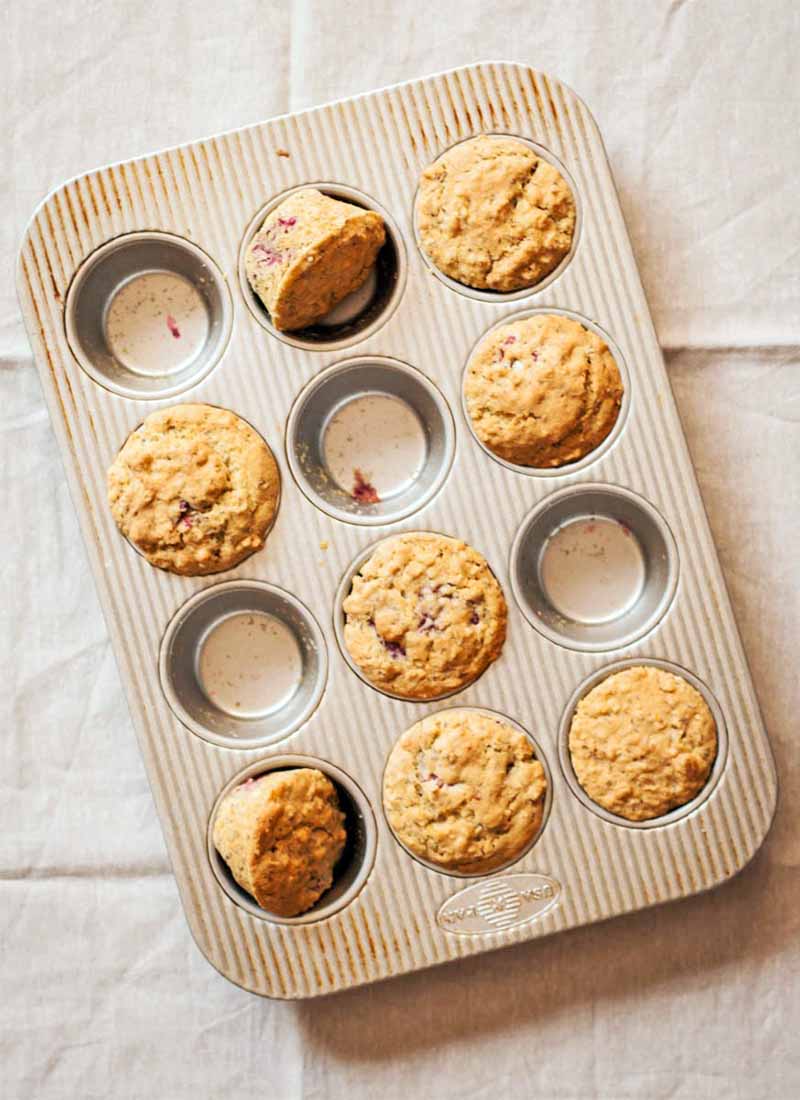 Top-down shot of a metal tin filled with eight just-baked muffins, two of which are tilted on their sides, on a wrinkled tan cloth background.